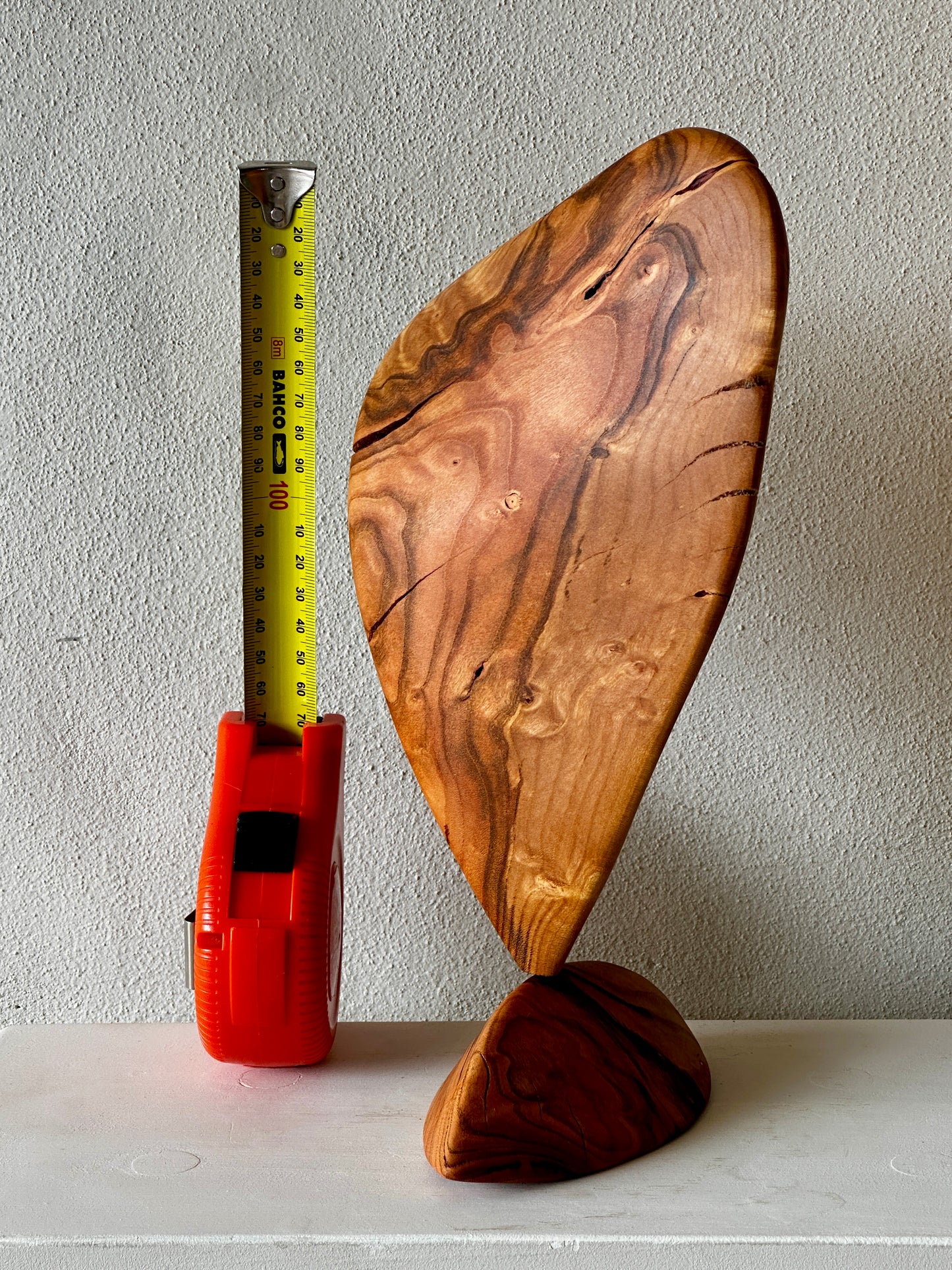 Wooden sculpture with a measuring tape for scale on a white surface.