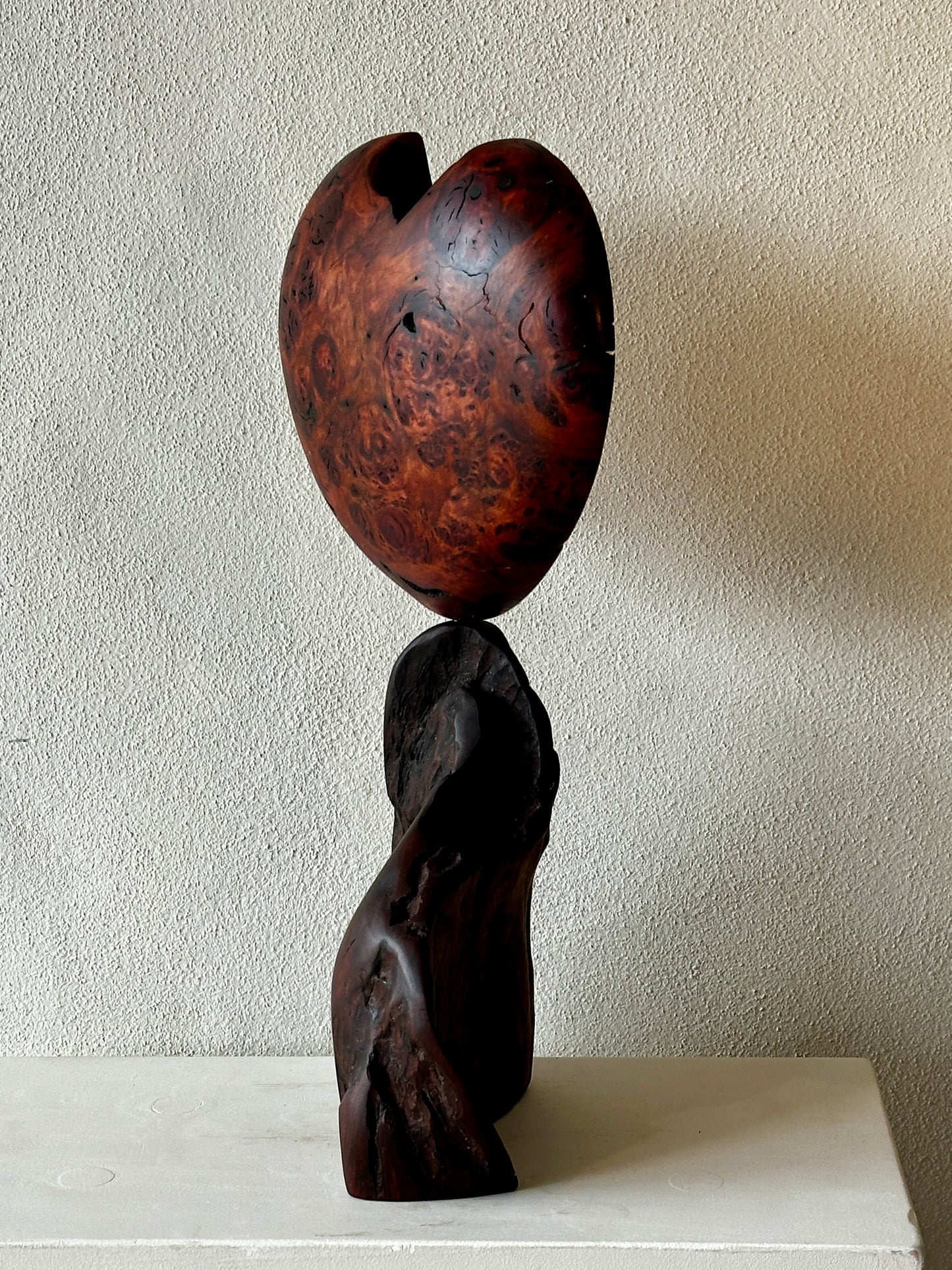 Wooden sculpture of a heart balanced on a twisted form against a plain wall.